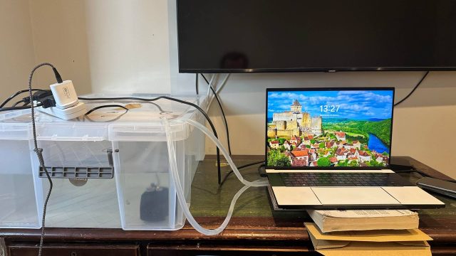 Laptop Razer Blade 16 đặt trên bàn, ống dẫn nước nối sang hộp nhựa chứa bơm