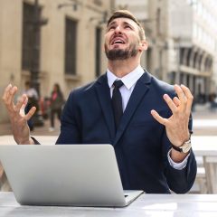 Người đàn ông ngồi trước laptop, giơ hai tay và ngửa mặt lên trời vì bực bội.