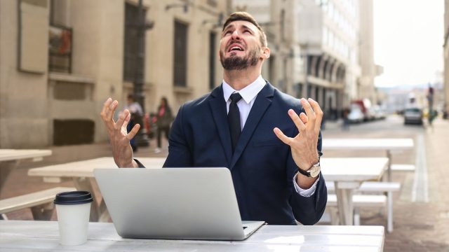 Người đàn ông ngồi trước laptop, giơ hai tay và ngửa mặt lên trời vì bực bội.