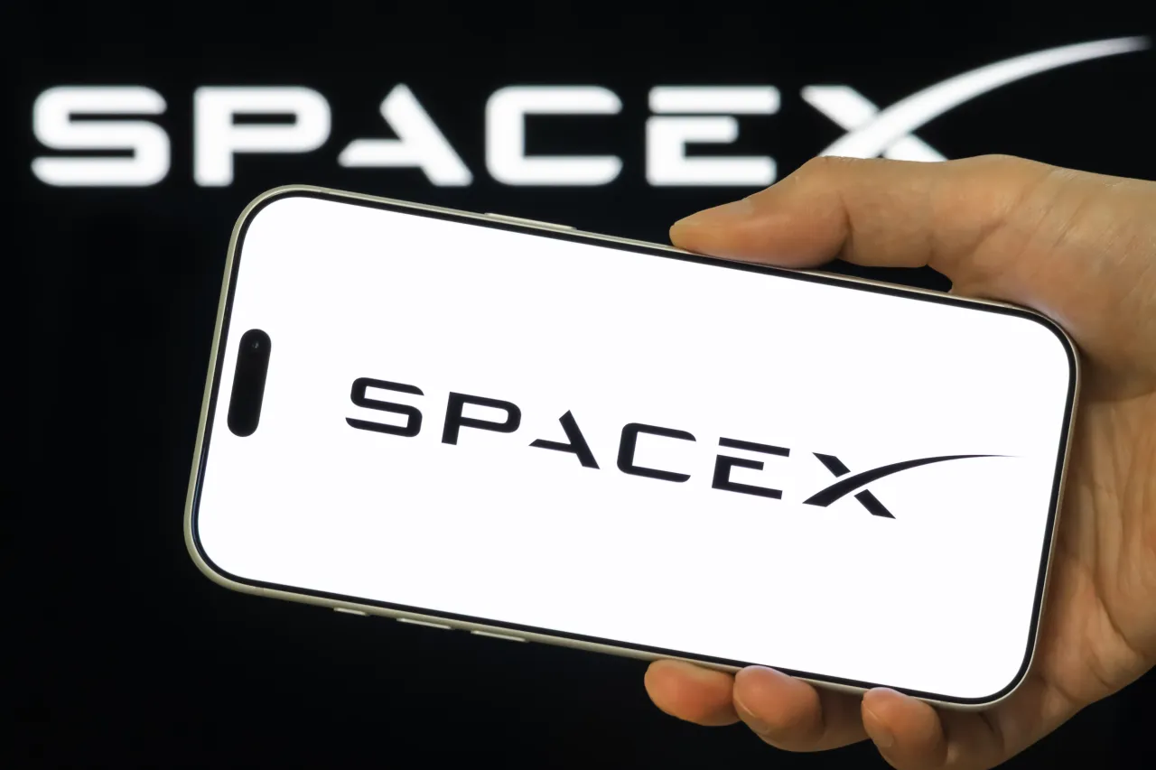 Bàn tay cầm điện thoại hiển thị logo SpaceX trên nền đen có chữ SpaceX