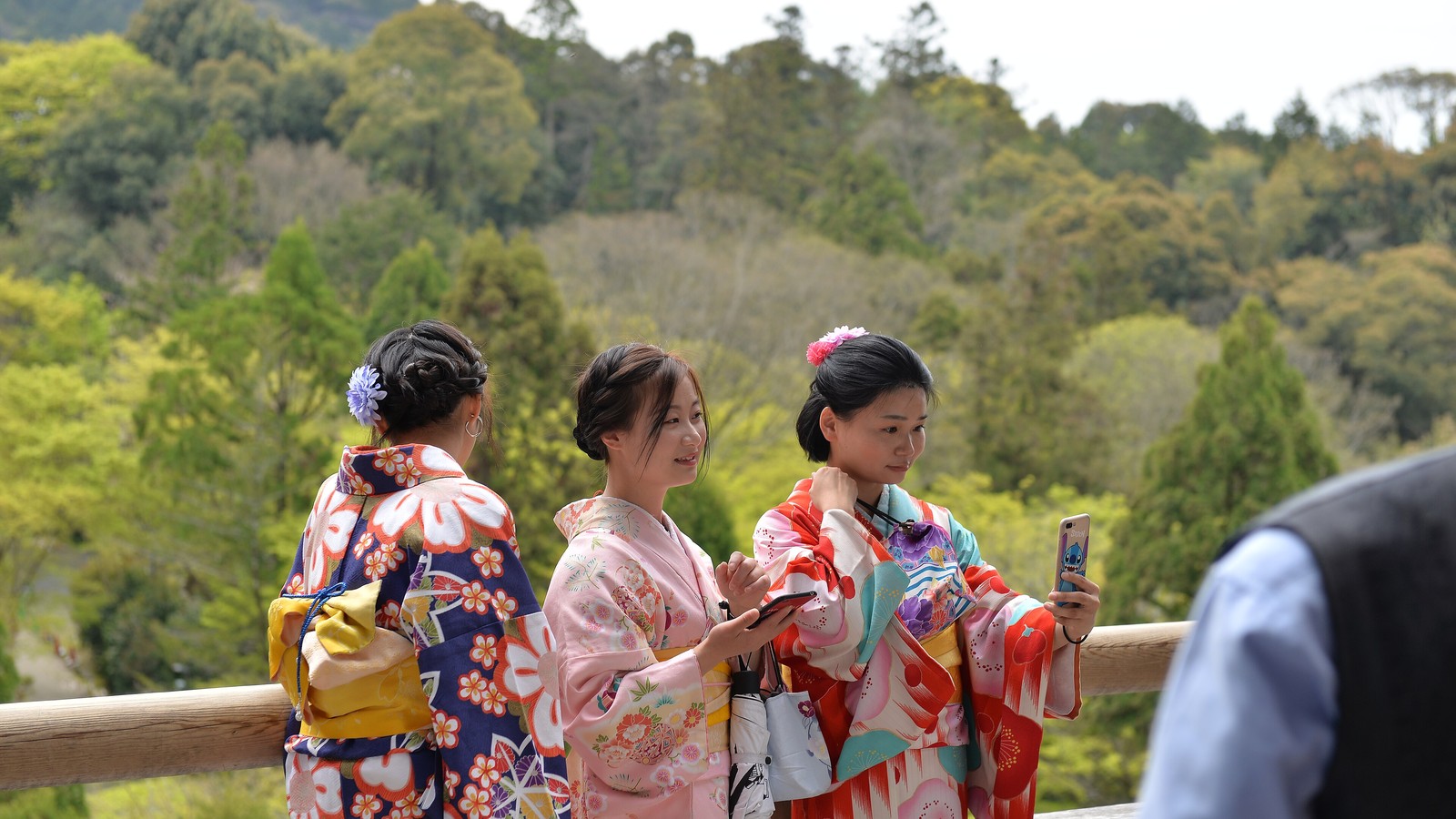 Three women in colourful kimonos looking at and holding up smartphones on a wooden balcony with green forested hills behind them
